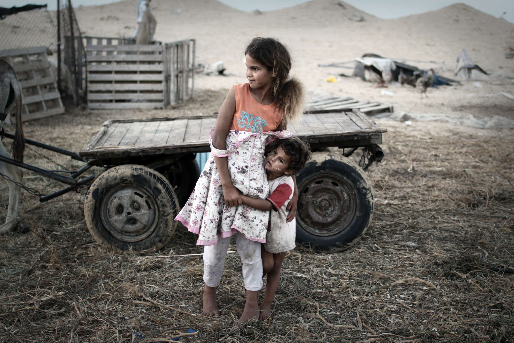 Two shoeless children embrace each other in a make shift camp site during the Gaza emergency. Occupied Palestinian Territory, 2013.
